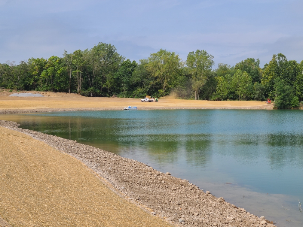 fresh straw surrounding a large retaining pond edged by a tree line; vehicles in background spreading straw and seed fresh straw surrounding a large retaining pond edged by a tree line; vehicles in background spreading straw and seed