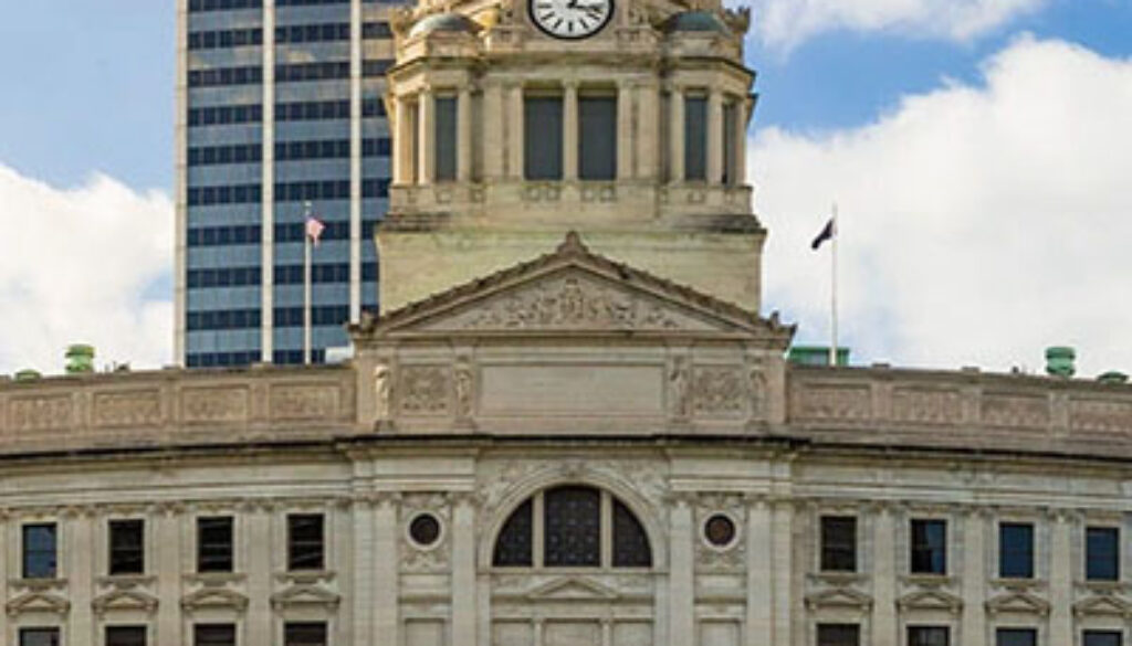 Front view of Fort Wayne courthouse with a clock tower and dome, with a modern skyscraper behind and a blue sky above
